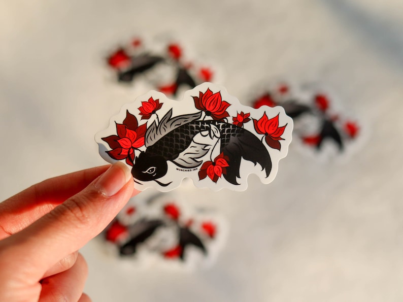Puede incluir: Una pegatina con un dise&ntilde;o de pez koi negro con flores de loto rojas. El pez tiene escamas detalladas y est&aacute; rodeado de flores rojas vibrantes. La pegatina tiene un borde blanco y una apariencia ligeramente transparente.