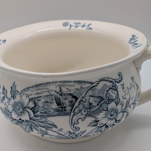 Könnte beinhalten: Antike weiße Porzellan-Nachttopf mit blauem Blumen- und Seelandschaftsdesign. Der Topf hat einen Griff und einen breiten Rand, mit detaillierten Illustrationen von Blumen und einem Segelschiff. Ein klassisches Stück Vintage-Badezimmerdekor.