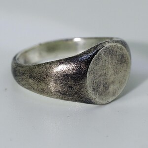 May include: A silver signet ring with a smooth, oval-shaped top. The ring has a brushed finish.