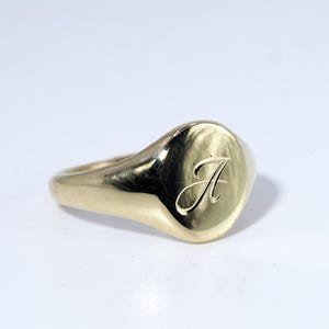 May include: A polished gold signet ring featuring a smooth, rounded face with a single, elegant script letter A engraved into the center. The band is solid and tapers slightly toward the back, showcasing a reflective, metallic finish throughout.