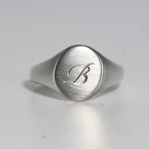 May include: A silver-toned signet ring with a brushed matte finish featuring an oval face. The center of the ring is engraved with a single, elegant cursive letter B. The band tapers slightly toward the back, maintaining a consistent metallic appearance.