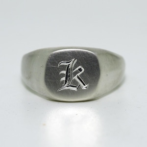 Op de afbeelding: Een zilverkleurige zegelring met een geborstelde, matte afwerking op het oppervlak. In het midden van de ring is een enkele, gestileerde letter K in een gotisch lettertype gegraveerd, wat een duidelijk contrast vormt met de gladde metalen band.