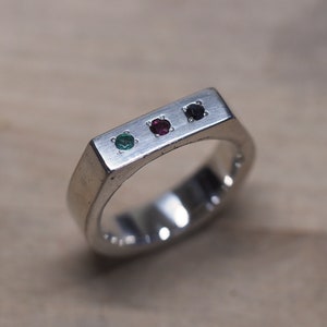 Może przedstawiać: Srebrny pierścionek z prostokątnym wierzchem, z trzema kamieniami szlachetnymi: zielonym, czerwonym i ciemnofioletowym. Pierścionek ma prosty, nowoczesny design i jest umieszczony na jasnej drewnianej powierzchni.