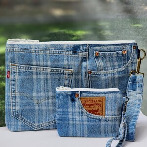 May include: A large and a small rectangular pouch made from light blue plaid denim fabric. Both pouches feature white zippers, visible denim pocket details, and copper-colored rivets. The smaller pouch includes a wrist strap and a brown leather label.