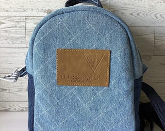 May include: A blue denim backpack with a brown leather patch that says "GUESS JEANS® LOS ANGELES" in white lettering. The backpack has a quilted design and a navy blue adjustable strap.