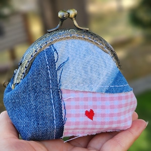 May include: A small coin purse featuring a patchwork design of blue denim fabric and light pink gingham fabric. The purse has a bronze-colored metal kiss-lock clasp with an ornate embossed pattern and a small red embroidered heart on the pink fabric.