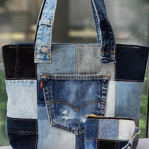 May include: A patchwork tote bag and matching wristlet pouch constructed from various shades of blue denim fabric. The tote features a prominent exterior pocket with a red tab and two shoulder straps. The pouch has a gold zipper and wrist strap.