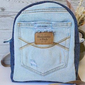 Puede incluir: Mochila de mezclilla azul claro con ribete azul oscuro y un bolsillo de mezclilla azul claro. La mochila presenta un parche de cuero marrón con el logotipo de Abercrombie & Fitch. La bolsa tiene correas ajustables y un cierre de metal.