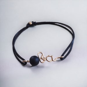 Peut inclure: Un bracelet en cordon noir avec une perle dorée et le mot "Bea" écrit en fil d'or.