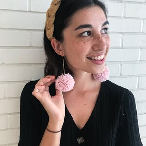 Può includere: Una donna indossa una fascia per capelli in velluto marrone e orecchini a pompon rosa. Indossa una camicetta nera con bottoni.