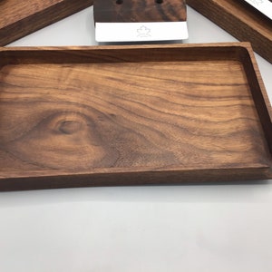 Può includere: Un vassoio rettangolare in legno con finitura marrone scuro. Il vassoio ha un bordo rialzato e una sezione scavata su un lato. La venatura del legno è visibile. Altri oggetti in legno sono sullo sfondo.