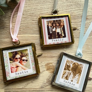 May include: Three small, square photo frames with gold and silver metal edges hang from ribbons. Each frame holds a photograph with the words "Family", "Bestie", and "Herbie" printed on the photos. The frames are displayed on a wooden surface.