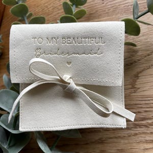 May include: A white, suede pouch with a white ribbon and the text "To my beautiful bridesmaid" embossed in gold.