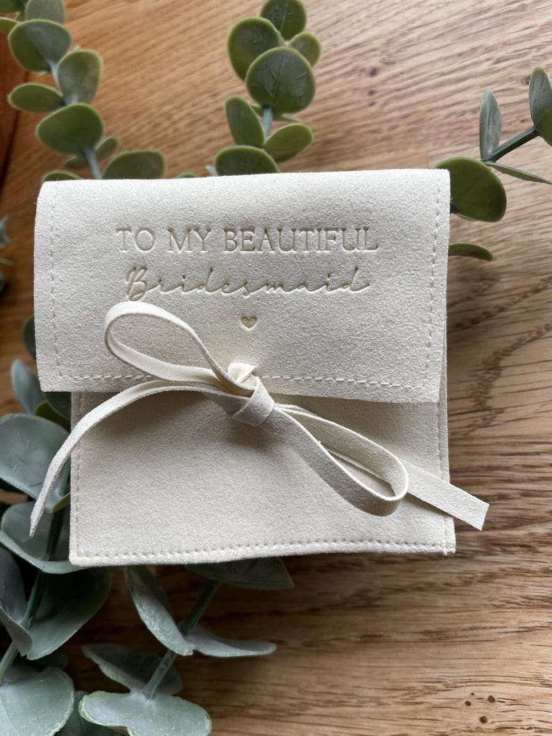 May include: A white, suede pouch with a white ribbon and the text "To my beautiful bridesmaid" embossed in gold.
