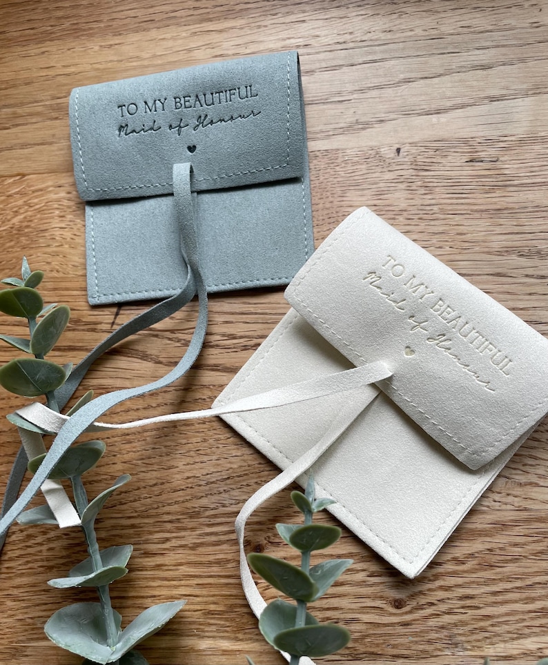 May include: Two small, fabric gift bags with a drawstring closure. The bags are made of a soft, suede-like material. The blue bag has the text "To my beautiful Maid of Honor" printed on the front. The white bag has the text "To my beautiful Maid of Honor" printed on the front.