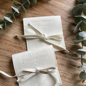 May include: Two white fabric pouches with a white ribbon tied around them. The pouches have the text "To my beautiful Mum" and "To my beautiful Flower girl" embossed on them.