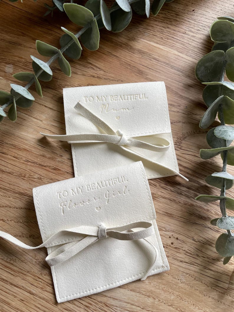 May include: Two white fabric pouches with a white ribbon tied around them. The pouches have the text "To my beautiful Mum" and "To my beautiful Flower girl" embossed on them.