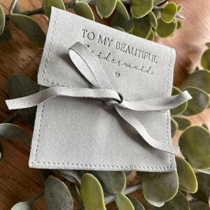 May include: A gray suede pouch with a white stitched border and a white ribbon tied in a bow. The pouch has the text "To my beautiful bridesmaid" embossed on the front.