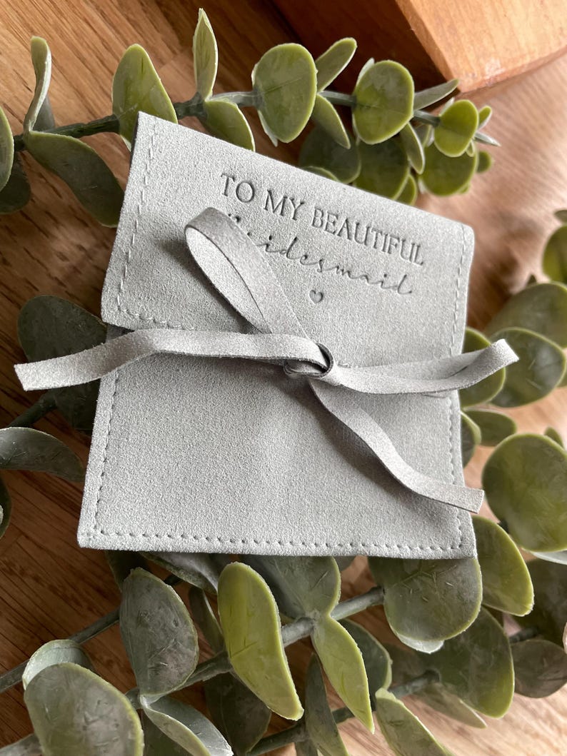 May include: A gray suede pouch with a white stitched border and a white ribbon tied in a bow. The pouch has the text "To my beautiful bridesmaid" embossed on the front.