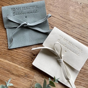 May include: Two small, fabric gift bags with a drawstring closure. The bags are made of a soft, suede-like material. One bag is light gray and the other is cream. Both bags have the text "To My Beautiful Bridesmaid" embossed on the front.