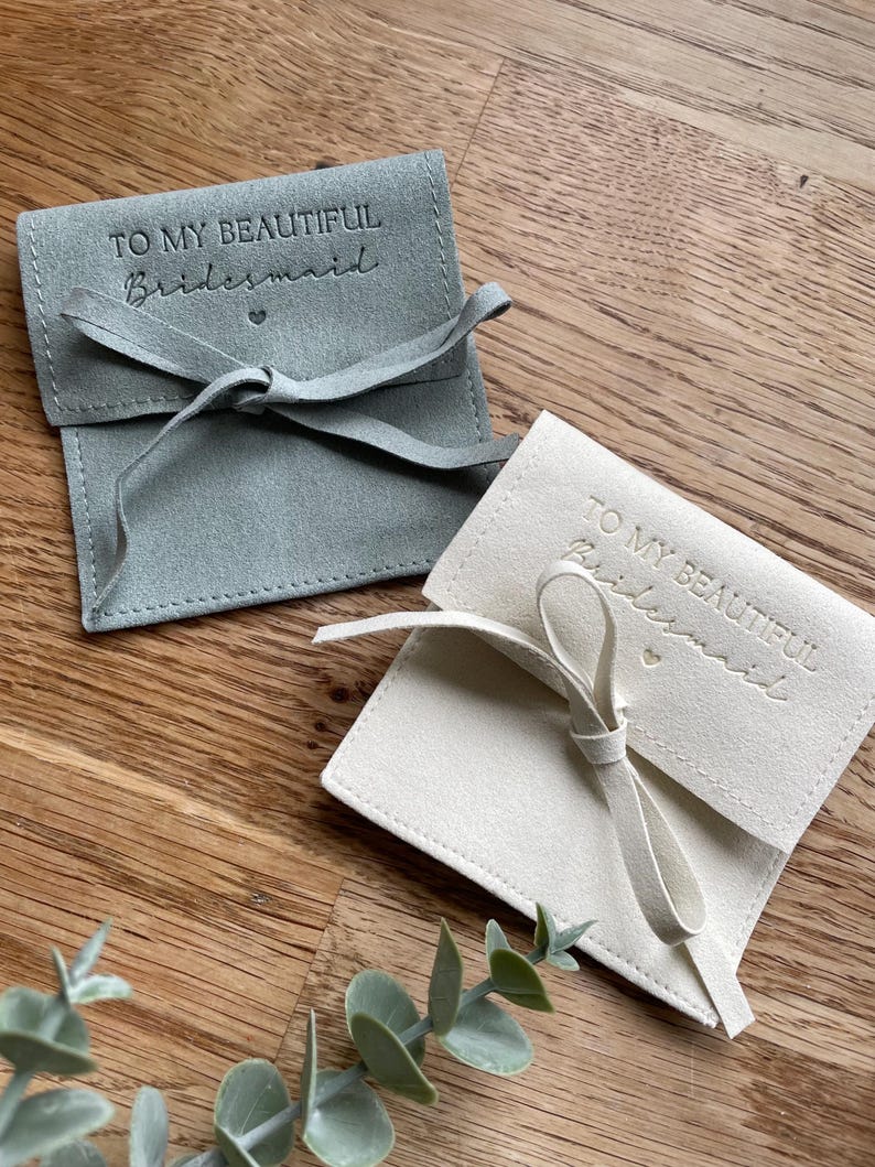May include: Two small, fabric gift bags with a drawstring closure. The bags are made of a soft, suede-like material. One bag is light gray and the other is cream. Both bags have the text "To My Beautiful Bridesmaid" embossed on the front.