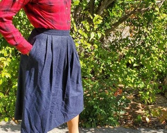 May include: A woman wearing a red and black plaid shirt and a dark blue pleated skirt. She is wearing brown ankle boots and is standing in front of a green bush.