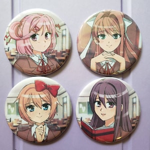 Puede incluir: Cuatro botones redondos con pasador que presentan personajes de anime del videojuego Doki Doki Literature Club. Los personajes son Monika, Sayori, Natsuki y Yuri. Cada botón tiene un color de fondo diferente y presenta el nombre del personaje en texto blanco.