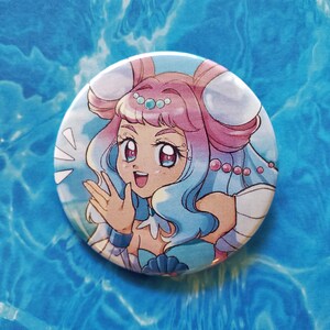 May include: A round button featuring a cartoon illustration of a girl with pink and blue hair, wearing a white and blue outfit with a seashell design. She is smiling and waving with her right hand.