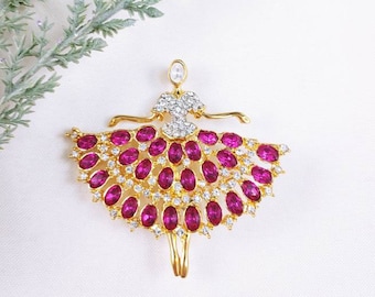 May include: A gold-toned brooch shaped like a ballerina, featuring a bodice encrusted with clear crystals and a wide, flared skirt decorated with numerous oval-shaped, vibrant magenta-pink stones and small clear accent crystals.