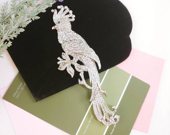May include: A silver-toned bird brooch encrusted with clear rhinestones, featuring a prominent crest and a small green stone for the eye. The bird is depicted perched on a branch with a long, flowing tail, showcasing intricate metalwork and sparkle.