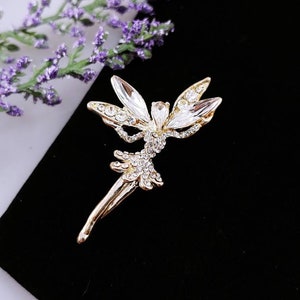 May include: A gold-toned fairy brooch with clear crystal embellishments. The fairy is depicted with wings outstretched and a flowing skirt.