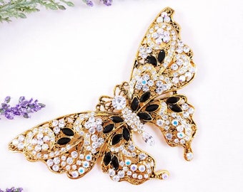 Puede incluir: Un broche de mariposa en tono dorado con intrincado trabajo de metal en filigrana, adornado con numerosas piedras transparentes brillantes y varias piedras negras alargadas y lisas dispuestas en un patrón decorativo en las alas.