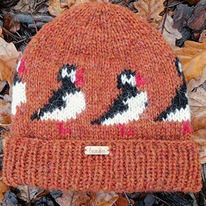 May include: A knitted orange beanie featuring a pattern of black, white, and red birds. The hat has a folded brim and a small wooden tag that reads "lumöss."