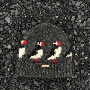 May include: A dark gray knit beanie featuring a repeating pattern of puffin birds in black, white, and red. The hat has a folded brim and a small, rectangular tag with text. The beanie is made of a textured, warm material.