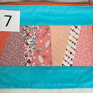 Peut inclure: Une pochette rectangulaire matelassée avec un dessus et un dessous bleu turquoise. Le centre présente un motif patchwork avec divers motifs floraux et géométriques en rose, pêche et orange. Le chiffre "7" est imprimé sur une étiquette blanche.