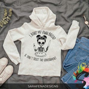 Könnte beinhalten: Ein weißer Hoodie mit einer schwarzen Grafik eines Skeletts, das Brot backt. Der Text auf dem Hoodie lautet "I bake my own bread & don't trust the government".