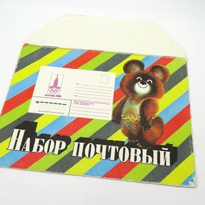 May include: Vintage envelope with a colorful striped pattern in red, yellow, blue, and green. Features a cartoon bear mascot with Olympic rings and Cyrillic text. Includes a postcard with the Moscow 1980 logo.