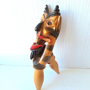 May include: A wooden nutcracker in the shape of a devil with a detailed face, horns, and a red tongue. The nutcracker has a light brown body with dark brown stripes and is standing on two wooden legs. The face is painted with tan, black, and red.