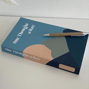 Peut inclure: Un journal intitulé "One Thought a Day" avec une couverture bleue et un design abstrait, un stylo, un vase beige avec de l'herbe de la pampa blanche et une bougie allumée dans un support bleu marine.
