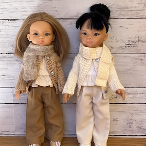 Puede incluir: Dos muñecas vestidas con conjuntos a juego. Una muñeca tiene el pelo rubio y la otra tiene el pelo negro. Ambas muñecas visten un top blanco, un suéter, una bufanda y pantalones. La muñeca rubia viste un conjunto marrón, y la otra un conjunto blanco.
