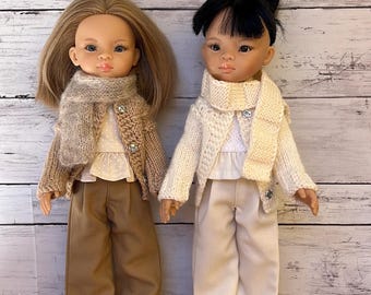 May include: Two dolls dressed in matching outfits. One doll has blonde hair and the other has black hair. Both dolls wear a white top, a sweater, a scarf, and pants. The doll with blonde hair wears a brown outfit, and the other doll wears a white outfit.