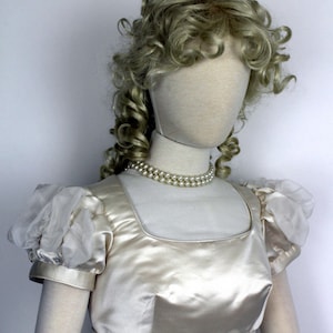 May include: A white satin dress with puffed sleeves and a pearl necklace. The dress has a square neckline and a fitted bodice.