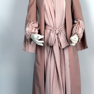 Op de afbeelding: Een lange, vloeiende roze robe met een witte kanten kraag en een bijpassende ceintuur die in de taille is geknoopt. De robe heeft lange, vloeiende mouwen met ruches aan de manchetten.