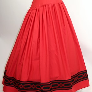 Puede incluir: Una falda roja vibrante con una cintura ancha y una silueta amplia y fluida. El dobladillo presenta una banda decorativa de bordado geométrico negro y rojo, agregando un detalle contrastante al diseño de la falda.