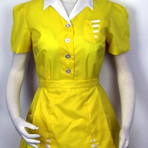 Puede incluir: Un uniforme amarillo con cuello blanco y un bolsillo blanco con rayas. El uniforme tiene falda y mangas cortas. El uniforme lo lleva un maniquí con una peluca rubia y un sombrero amarillo.