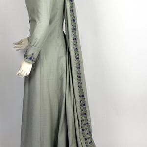 Może przedstawiać: Długa, szara sukienka z trenem. Sukienka ma purpurowo-zieloną haftowaną lamówkę wokół dołu i trenu.