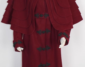 Può includere: Un lungo cappotto di lana color borgogna con un ampio mantello a più strati. Il cappotto è rifinito con pizzo nero e bottoni decorativi.