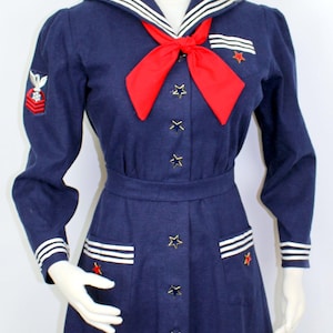 May include: A navy blue dress with a sailor collar and white stripes. The dress has a red bow tie and gold star buttons. The dress has two pockets with white stripes and a gold star on each pocket.