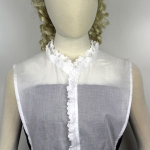 May include: White cotton lace collar with a ruffled edge. The collar is sleeveless and has a button closure in the front.