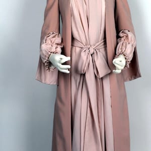 May include: A long, flowing pink robe with a white lace collar and a matching sash tied at the waist. The robe has long, flowing sleeves with ruffles at the cuffs.